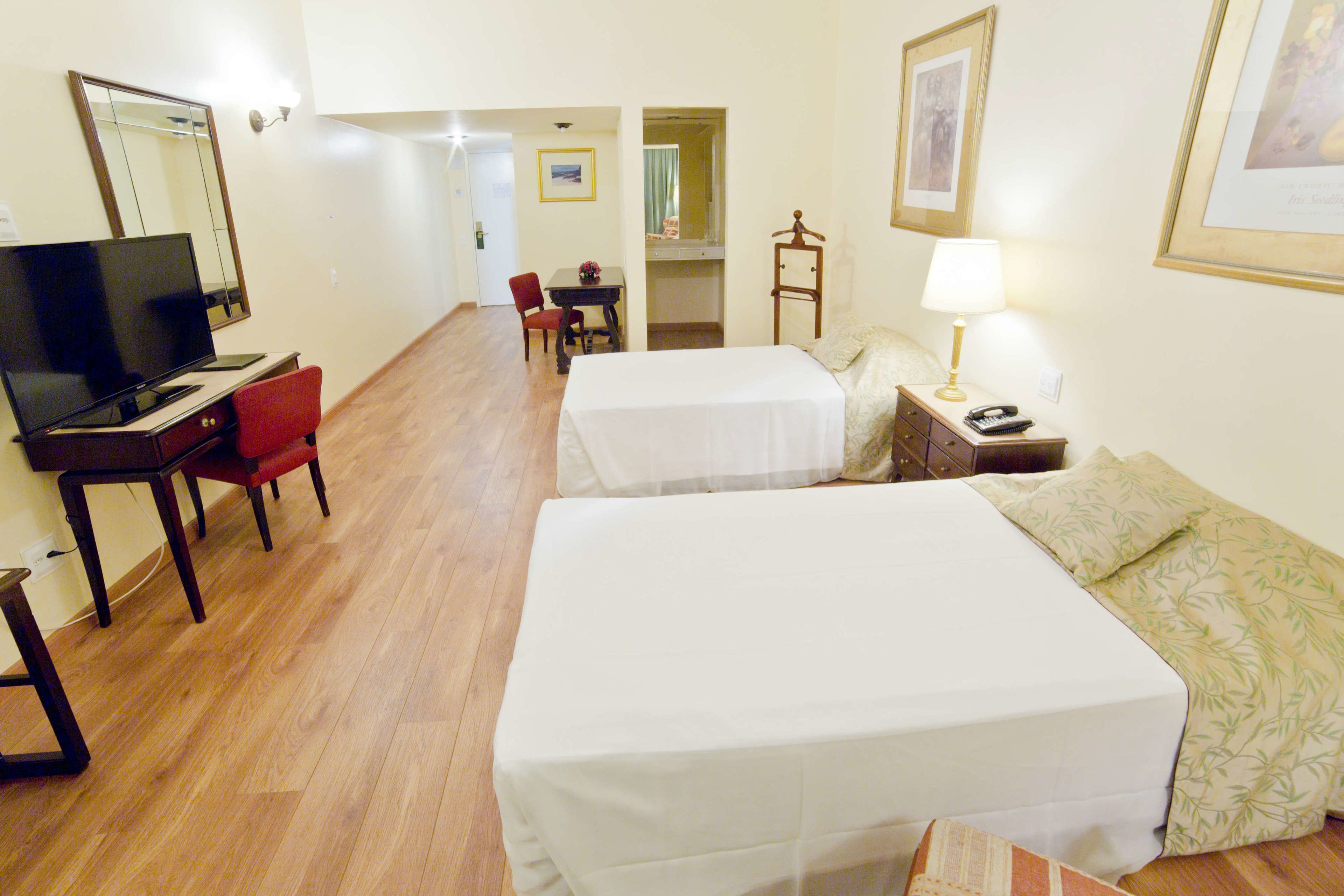 Quarto de hotel com duas camas, uma secretária com computador, uma cadeira, um abajur e um espelho.