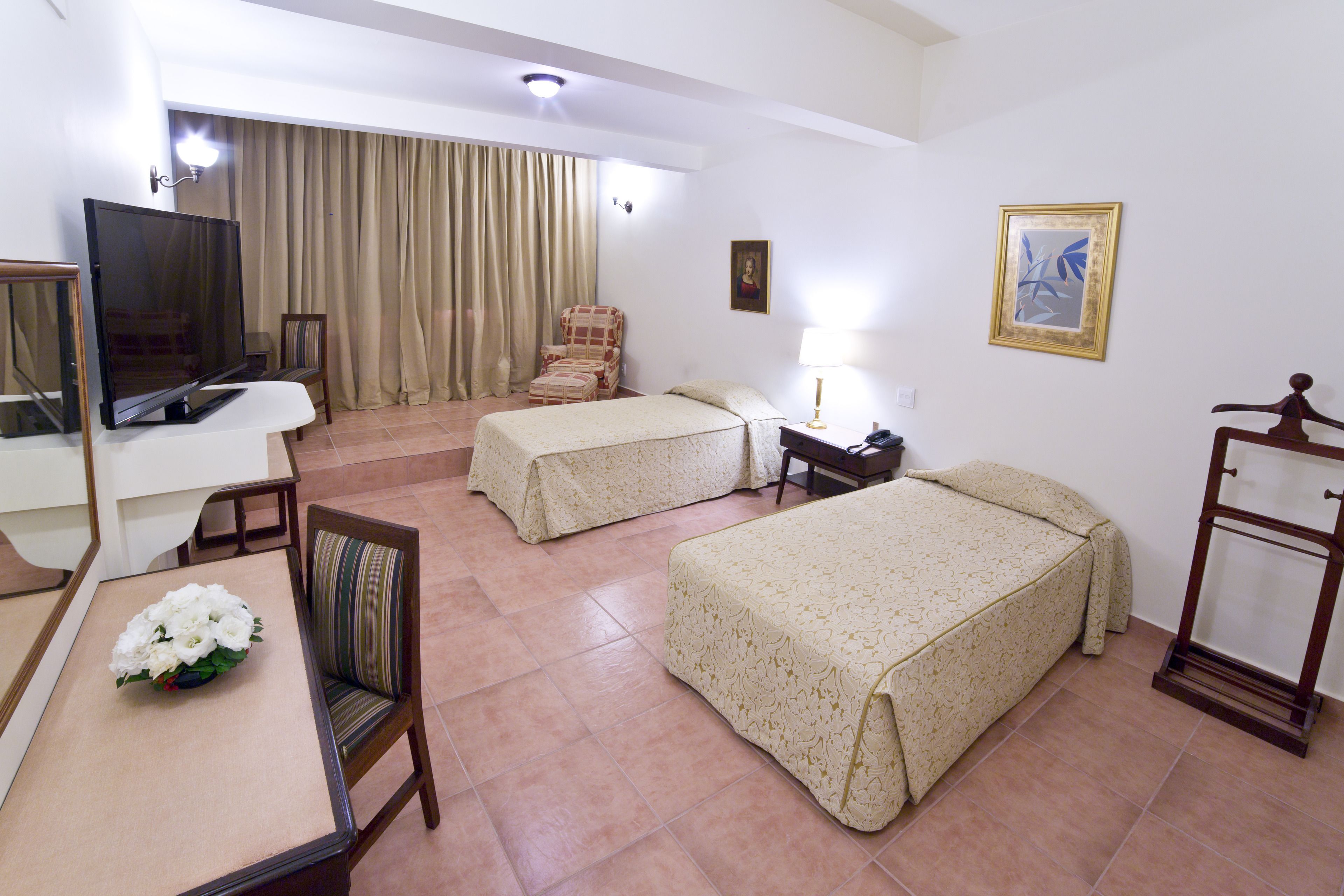 Habitación de hotel con dos camas, un escritorio con televisor, una silla, una lámpara y un cuadro en la pared.