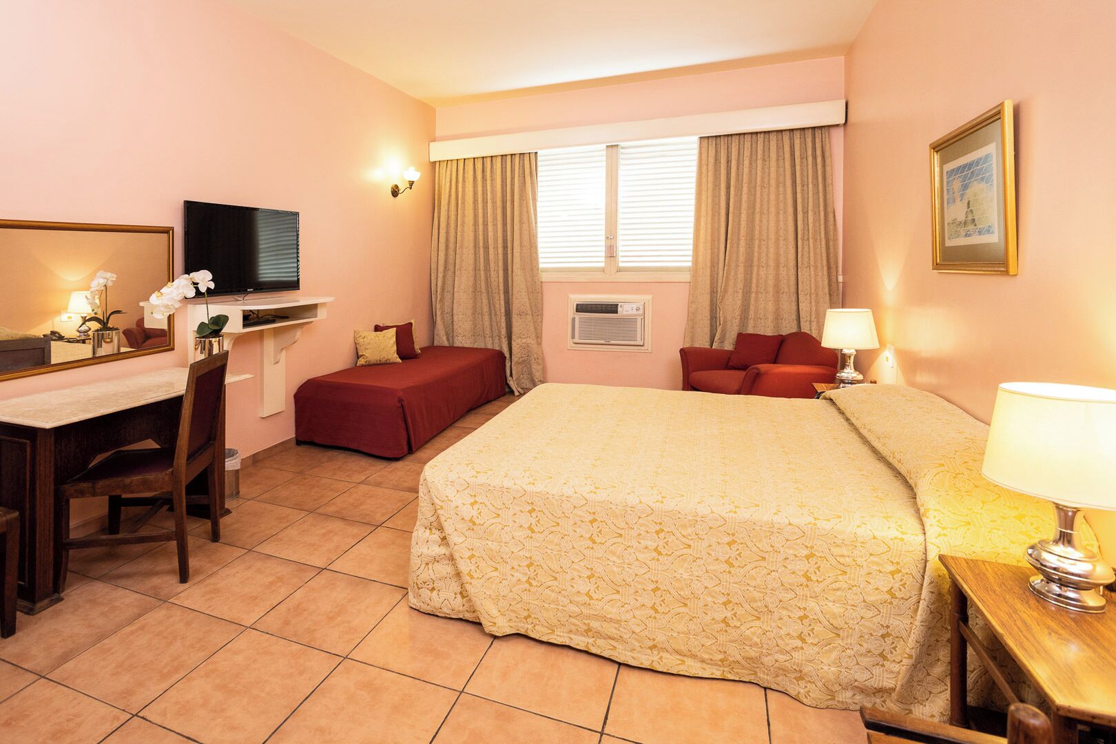 A hotel room with a bed, a desk with a chair, a TV, a window with curtains, and a framed picture on the wall.