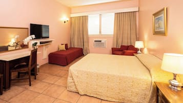 A hotel room with a bed, a desk with a chair, a TV, a window with curtains, and a framed picture on the wall.
