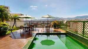 Zona de piscina en la azotea con vistas a la ciudad, rodeada por una valla verde y con mobiliario de exterior.