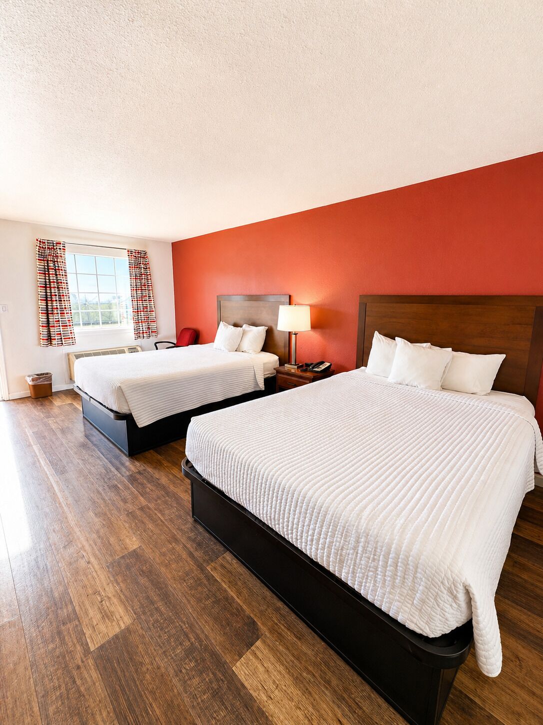 Une chambre d’hôtel avec deux lits, une fenêtre avec des rideaux, une table de chevet avec une lampe et un plancher en bois.