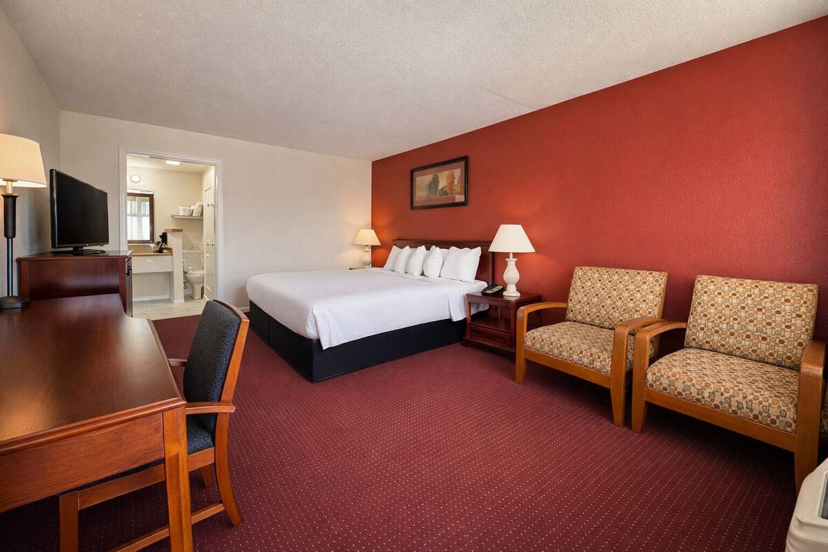 A hotel room with a bed, two armchairs, a desk, a television, and a painting on the wall.