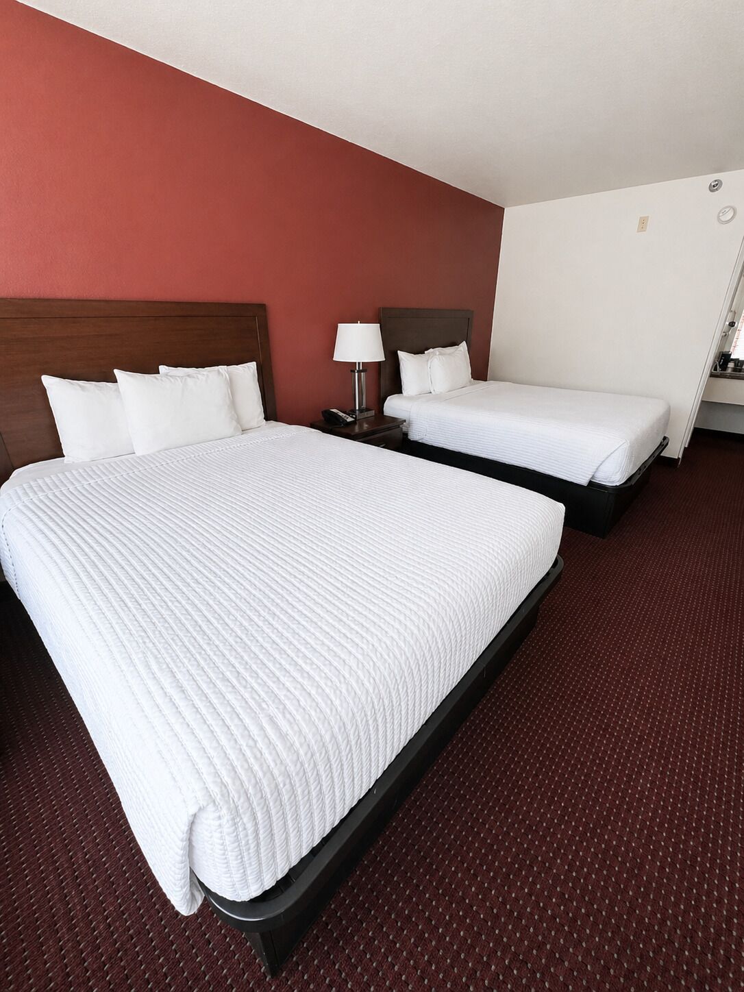 Quarto de hotel com duas camas, cabeceira de madeira, mesa de cabeceira com luminária e piso coberto por carpete vermelho.