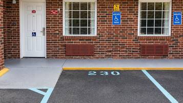 Un bâtiment à la façade en briques, doté de deux fenêtres, d’une porte avec un panneau indiquant une place de stationnement réservée aux personnes handicapées, et d’une place de stationnement marquée du numéro « 230 ».