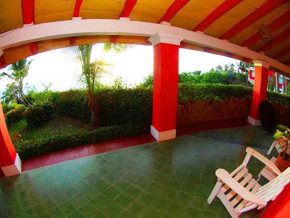 A covered outdoor area with a tiled floor, a white chair, and a view of a landscaped garden with a palm tree and red flowers. - Hotel Los Flamingos (Acapulco)