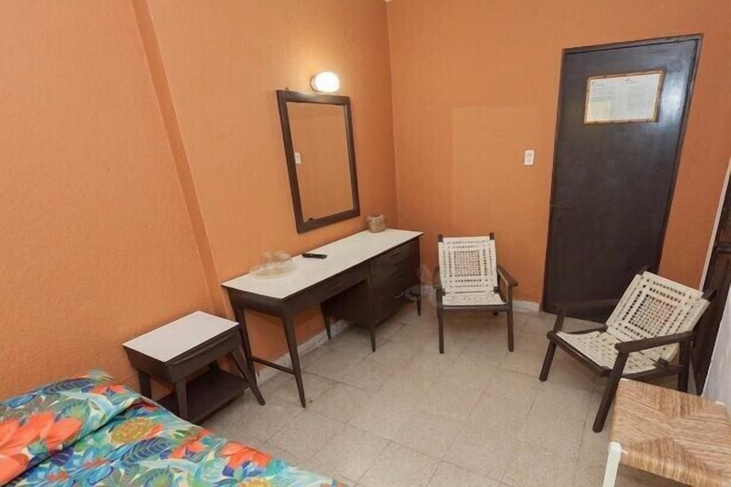 Una habitación pequeña y funcional con una cama individual, un escritorio, dos sillas y una puerta.