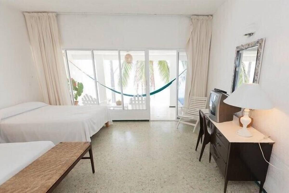 A hotel room with a bed, a desk with a lamp, a TV, and a view of palm trees through the window.