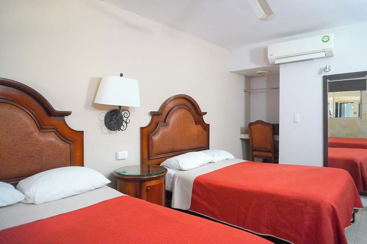 Habitación de hotel con dos camas, cada una con cabecero de madera y cubrecamas rojos. Se observa una lámpara de pared, una mesita de noche y un equipo de aire acondicionado.