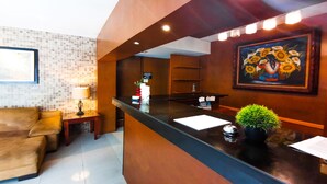 A hotel reception area with a wooden reception desk, a couch, a lamp, a framed painting, and a potted plant. - Hotel Colonial Cancun (Cancun)
