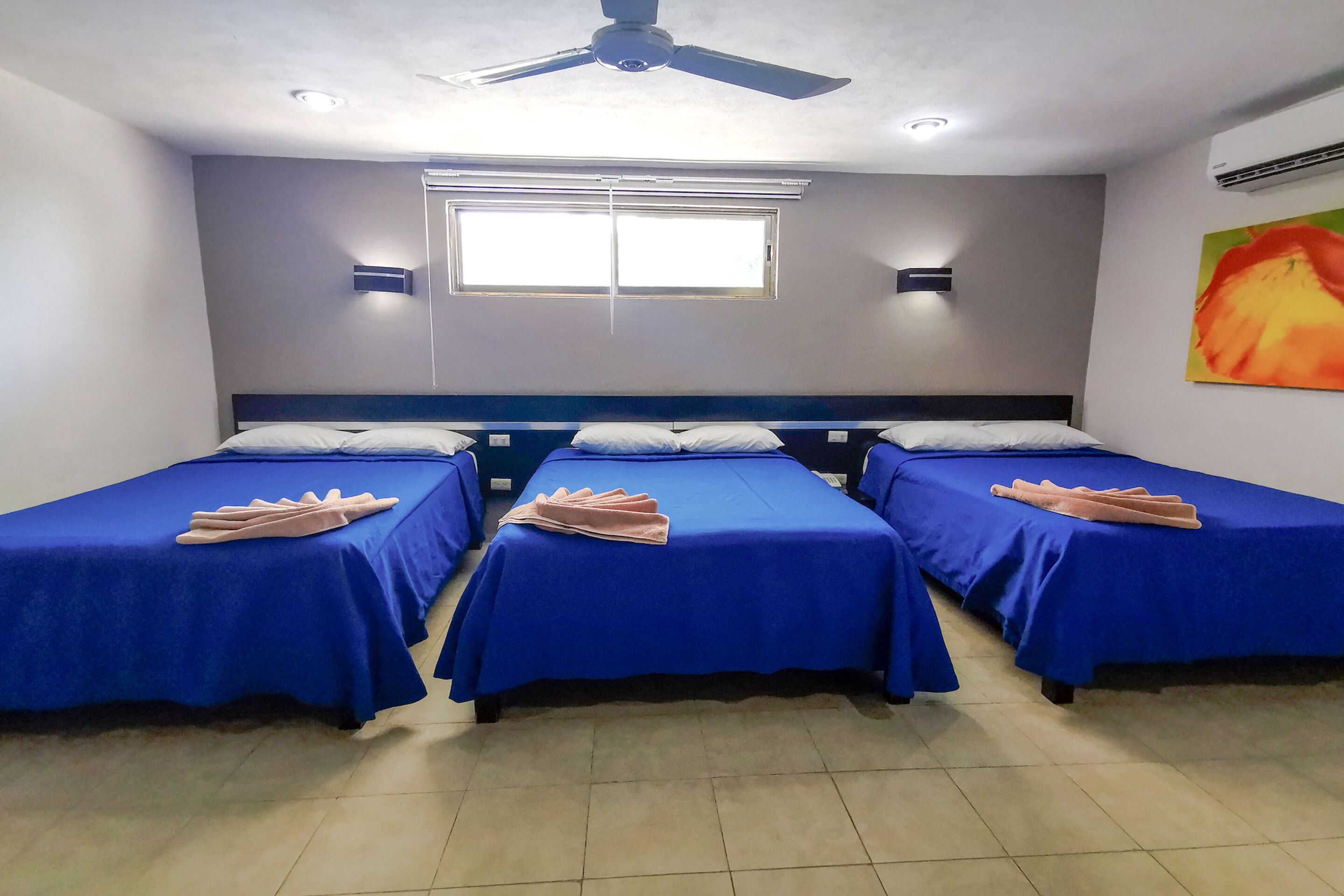 Quarto de hotel com três camas de solteiro, cada uma com colcha azul e toalha rosa. Há um ventilador de teto, uma janela com persianas e um ar condicionado fixado na parede.