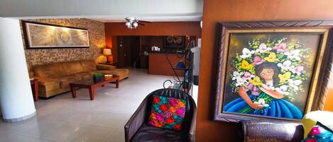 A spacious living room with a sofa, a chair with a colorful cushion, a small table with a plant, and a painting of a woman with flowers.