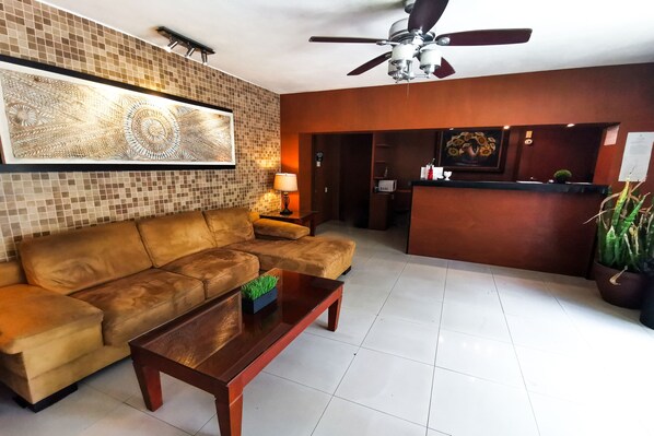 A hotel lobby with a brown leather sofa, a wooden coffee table, a ceiling fan, a reception desk, and a large wall art piece. - Hotel Colonial Cancun (Cancun)