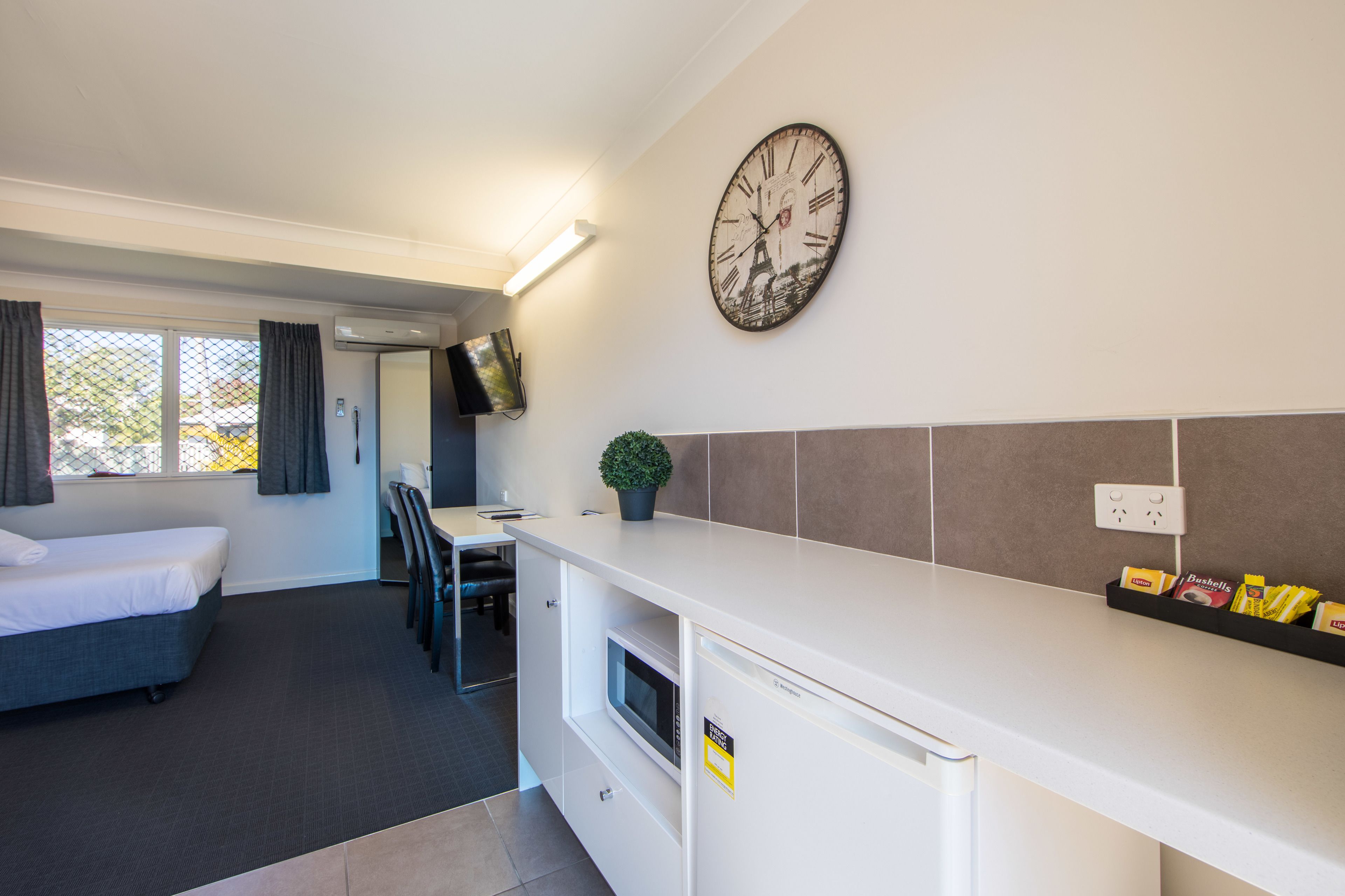 A compact hotel room with a kitchenette, a bed, a small dining area, and a large wall clock.
