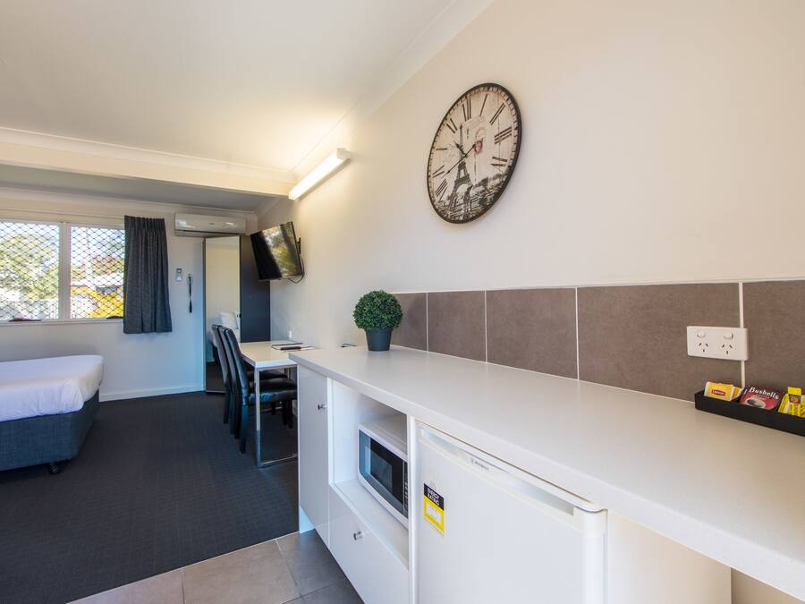 A compact hotel room with a kitchenette, a bed, a small dining area, and a large wall clock.