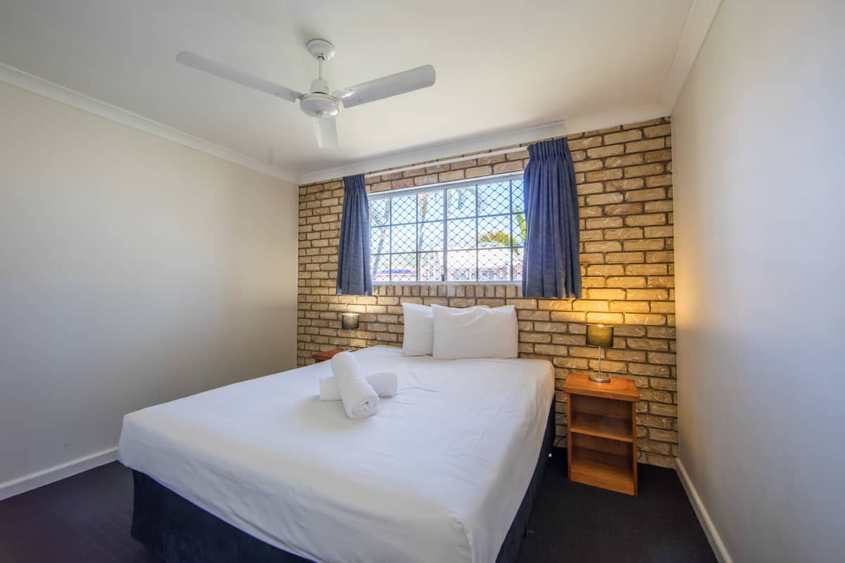 Habitación de hotel con cama, ventilador de techo, pared de ladrillo y ventana con cortinas.