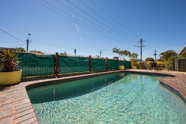 Una piscina cristalina con borde de ladrillo, rodeada por una valla verde y plantas en macetas.