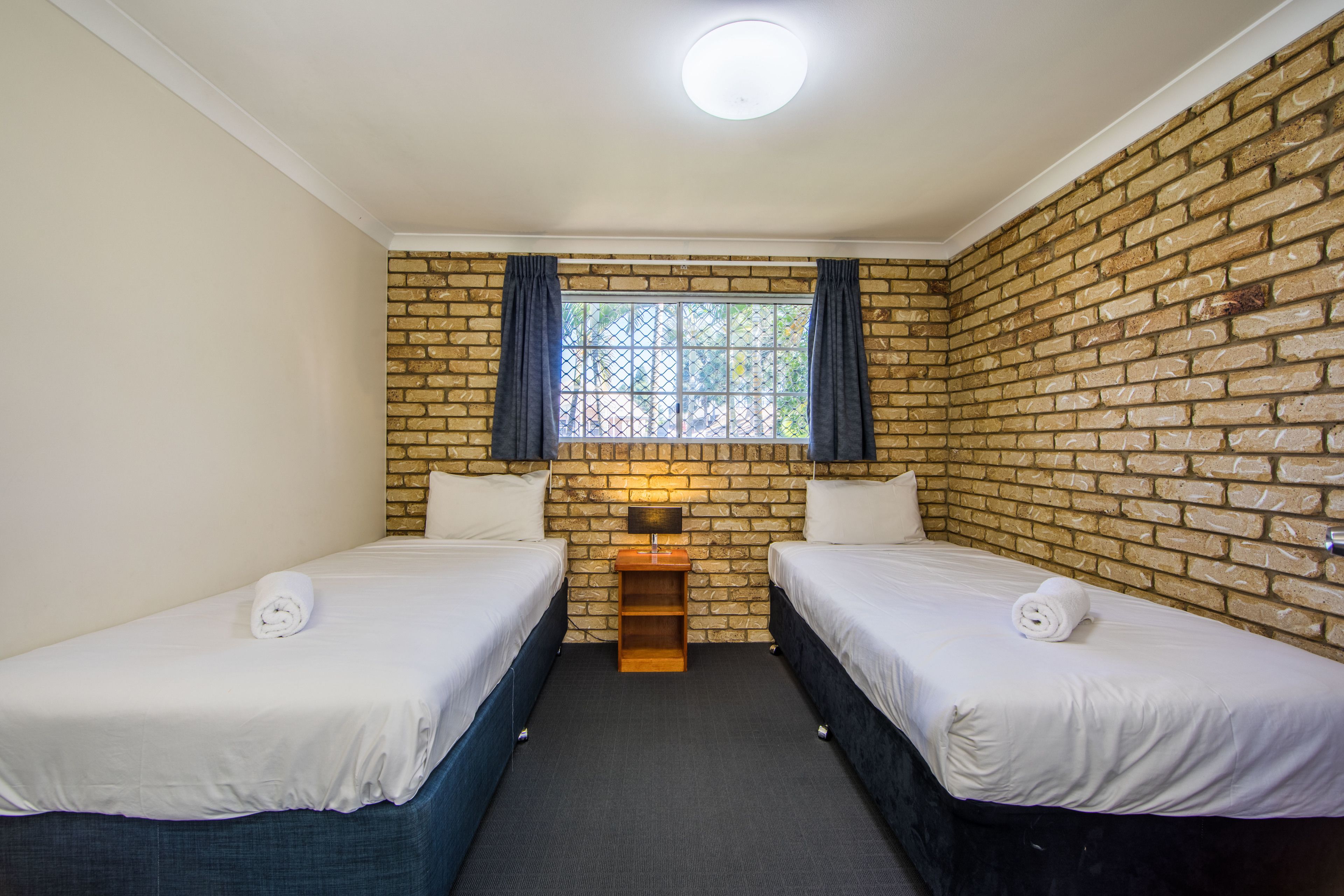 A room with two beds, a brick wall, a window with curtains, and a small table with a lamp.