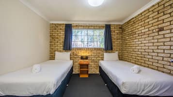 A room with two beds, a brick wall, a window with curtains, and a small table with a lamp.