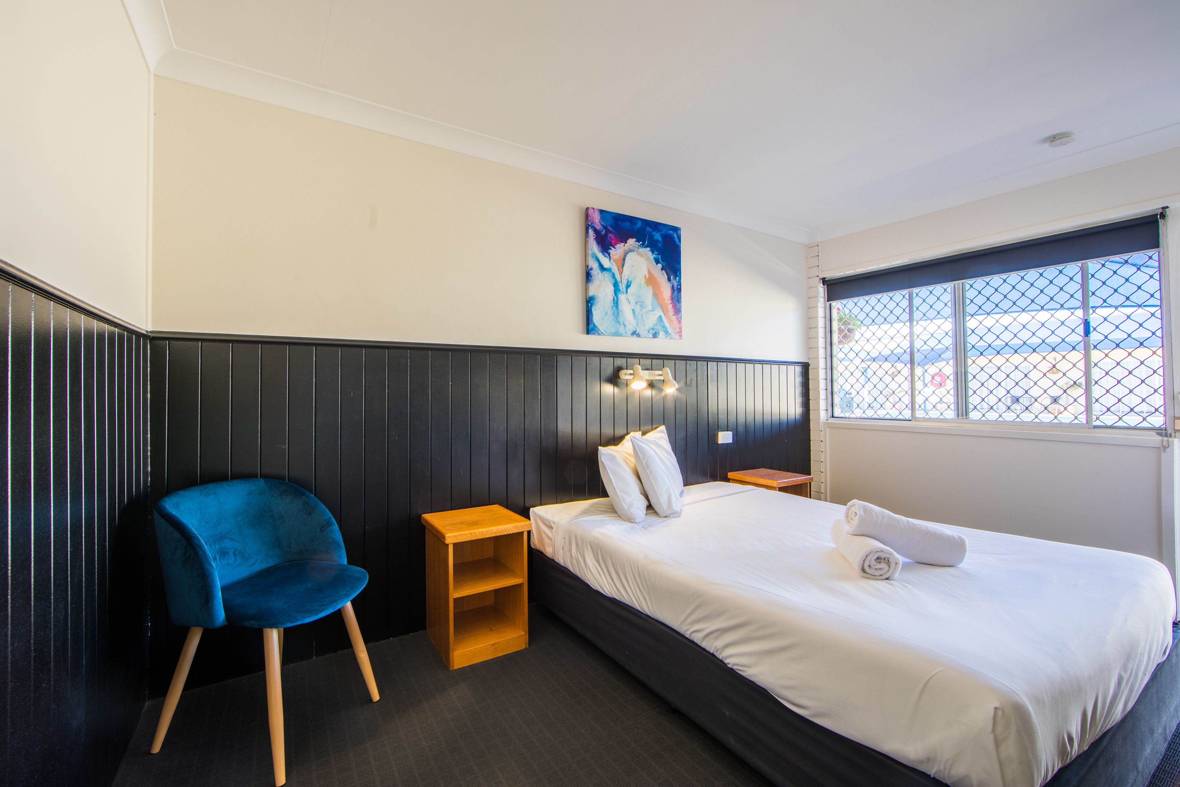A hotel room with a bed, a blue chair, a small wooden nightstand, a painting on the wall, and a window with a view of the outside.