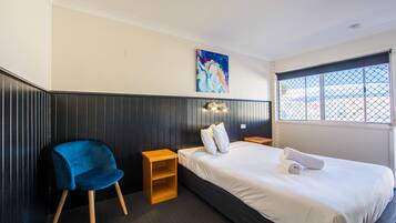 A hotel room with a bed, a blue chair, a small wooden nightstand, a painting on the wall, and a window with a view of the outside.