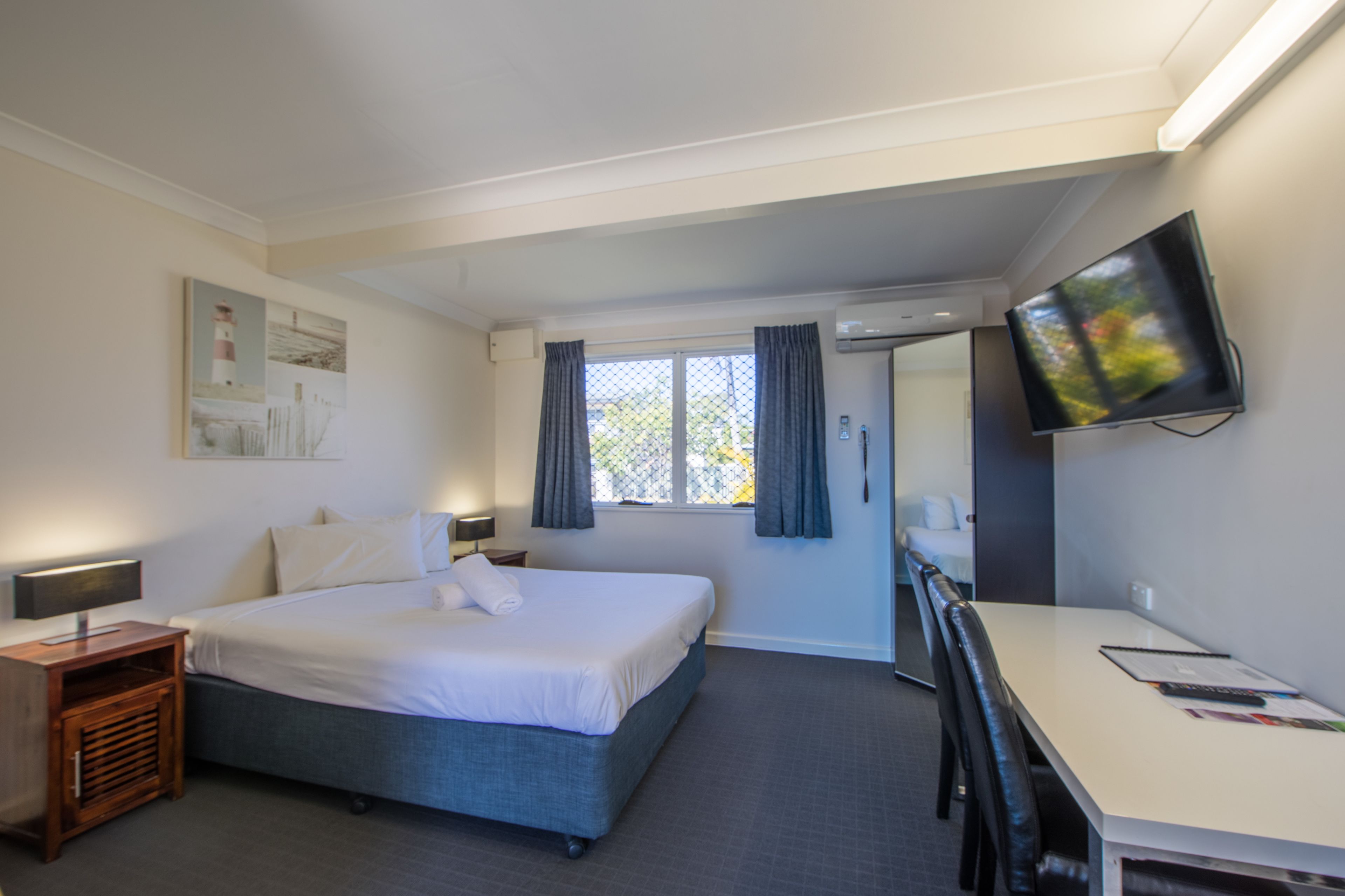 A hotel room with a bed, bedside table, desk, chair, TV, window with curtains, and artwork on the wall.