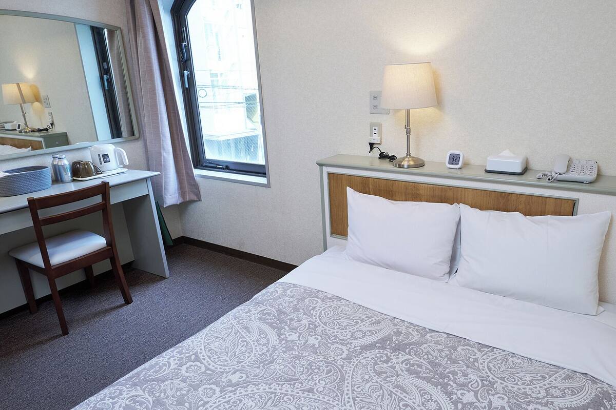 A hotel room with a bed, a desk with a chair, a mirror, a window with curtains, a bedside table with a lamp, and a phone.