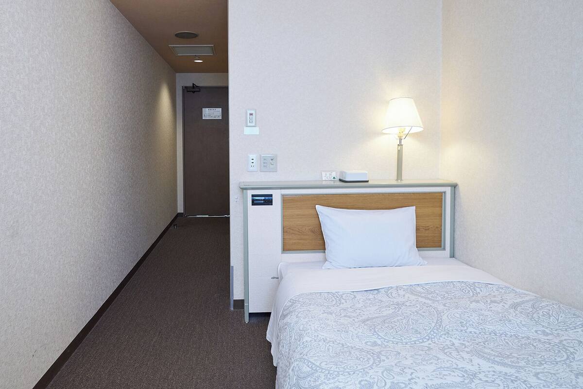 A hotel room with a bed, a bedside lamp, a nightstand, a wall clock, and a door leading to another room.