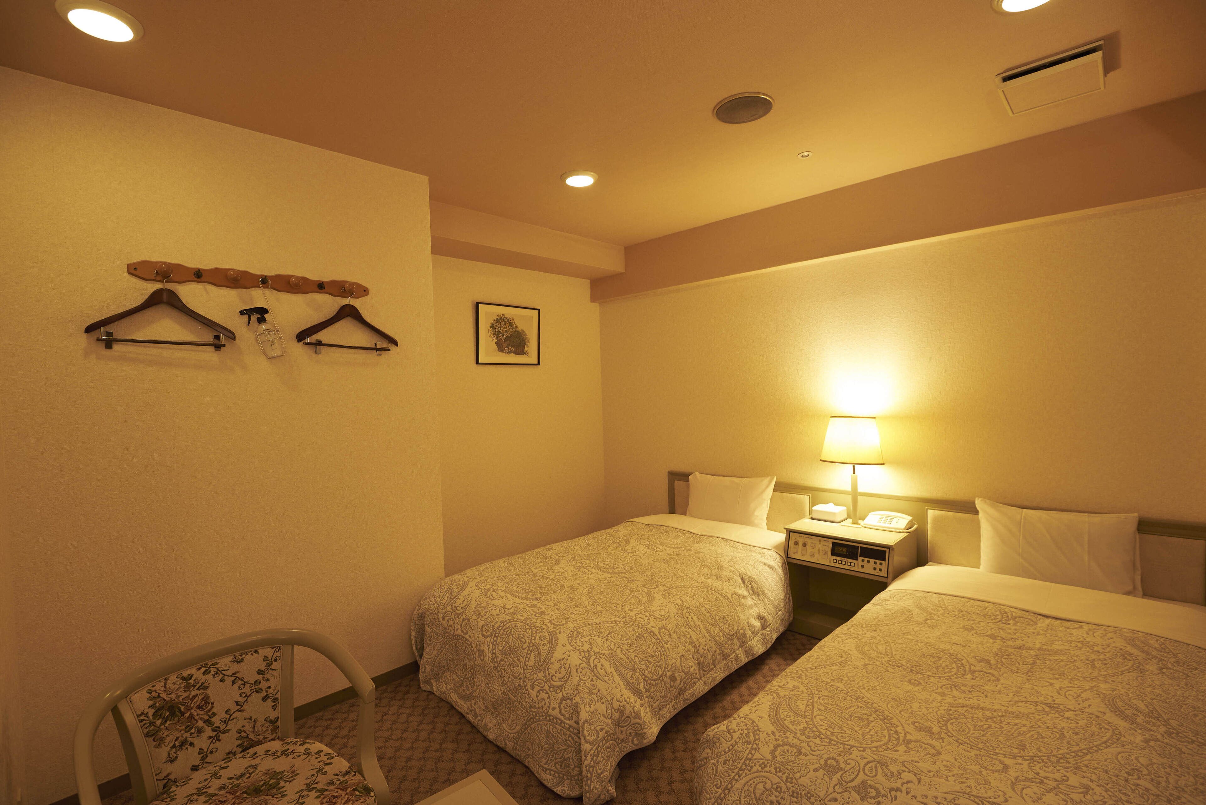 A hotel room with two beds, a chair, a bedside table with a lamp, and a wall-mounted coat rack.