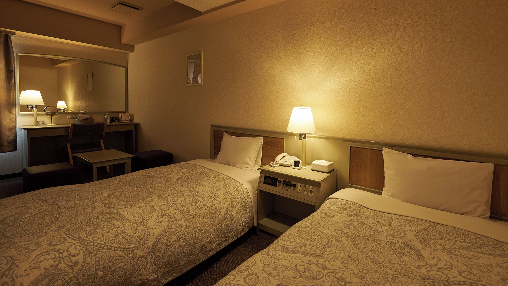 Quarto de hotel com duas camas, uma escrivaninha com abajur, um telefone e um espelho.