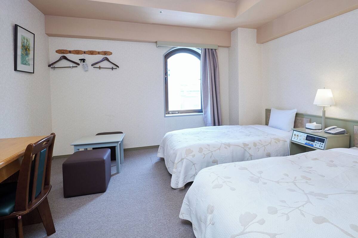 A hotel room with two beds, a desk, a chair, a small table, and a window with curtains.