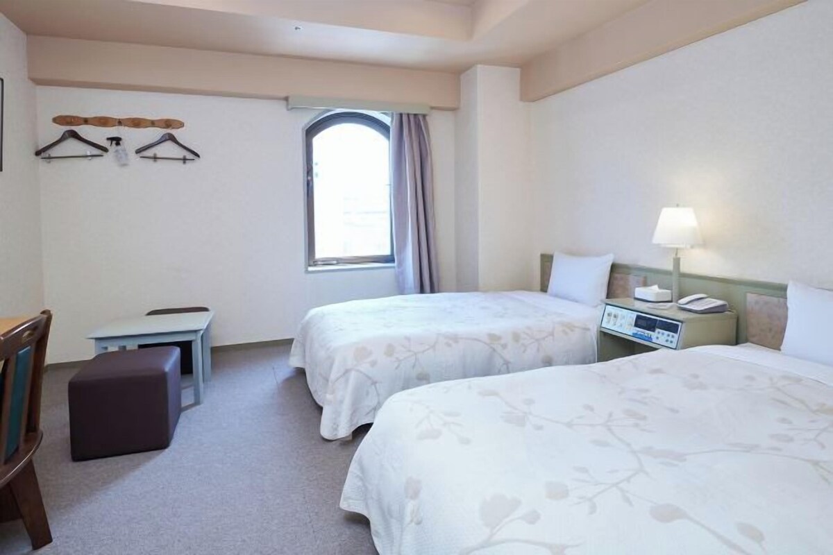 A hotel room with two beds, a desk, a chair, a small table, a window, and a nightstand with a lamp.