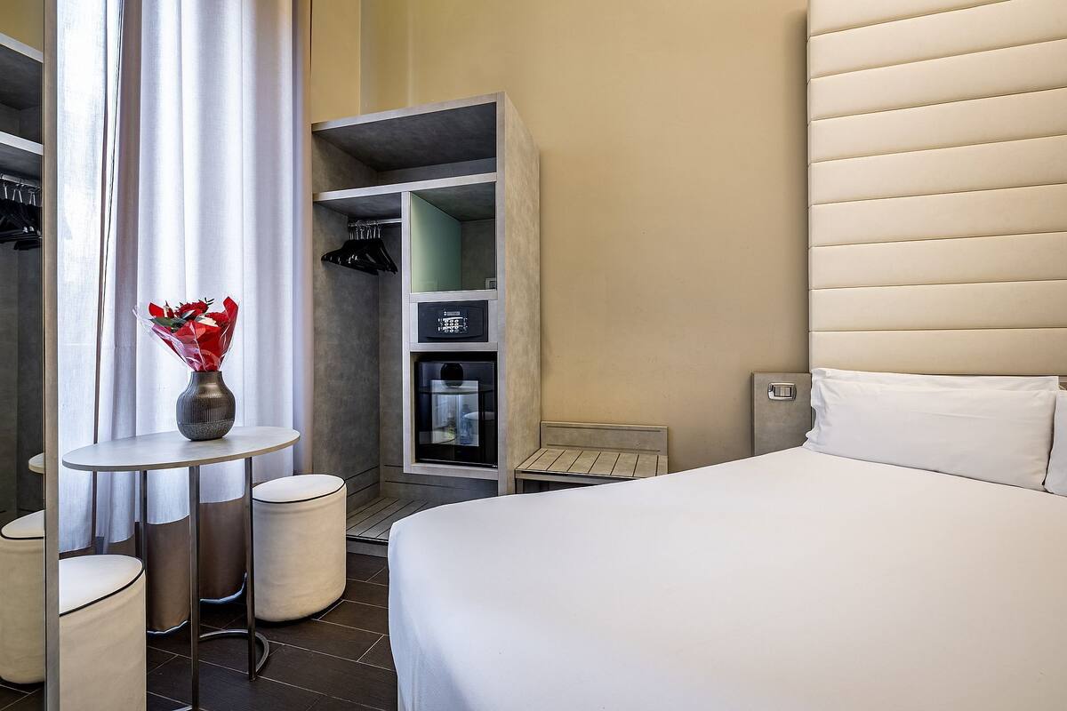 A hotel room with a bed, a vanity with a mirror, a small table with a vase, a closet, and a minibar.