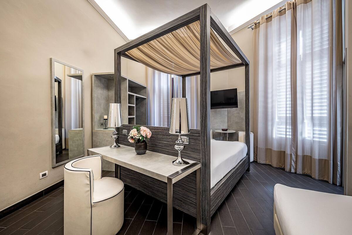 A hotel room with a four-poster bed, a desk with a lamp, a chair, a large mirror, and a television.