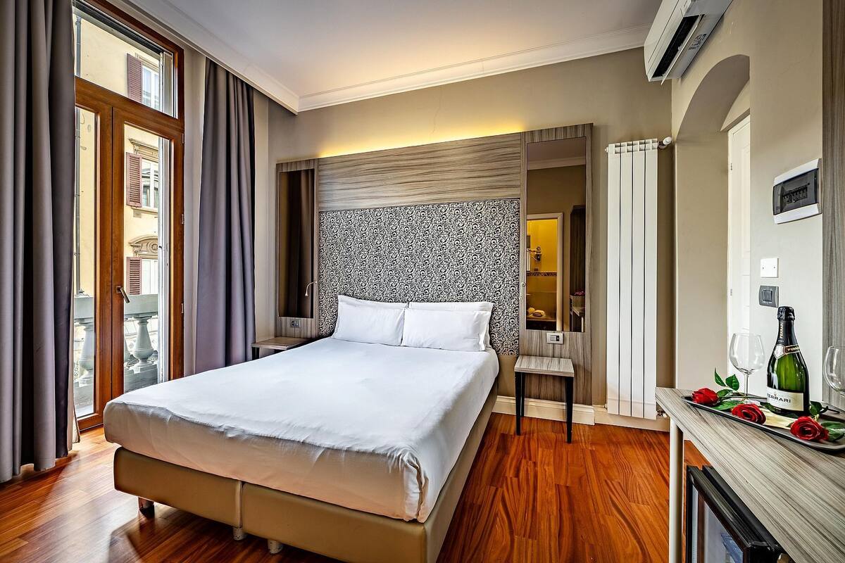A hotel room with a bed, a vanity with a mirror, a small table with a bottle and glasses, and a view of the outside through a window.