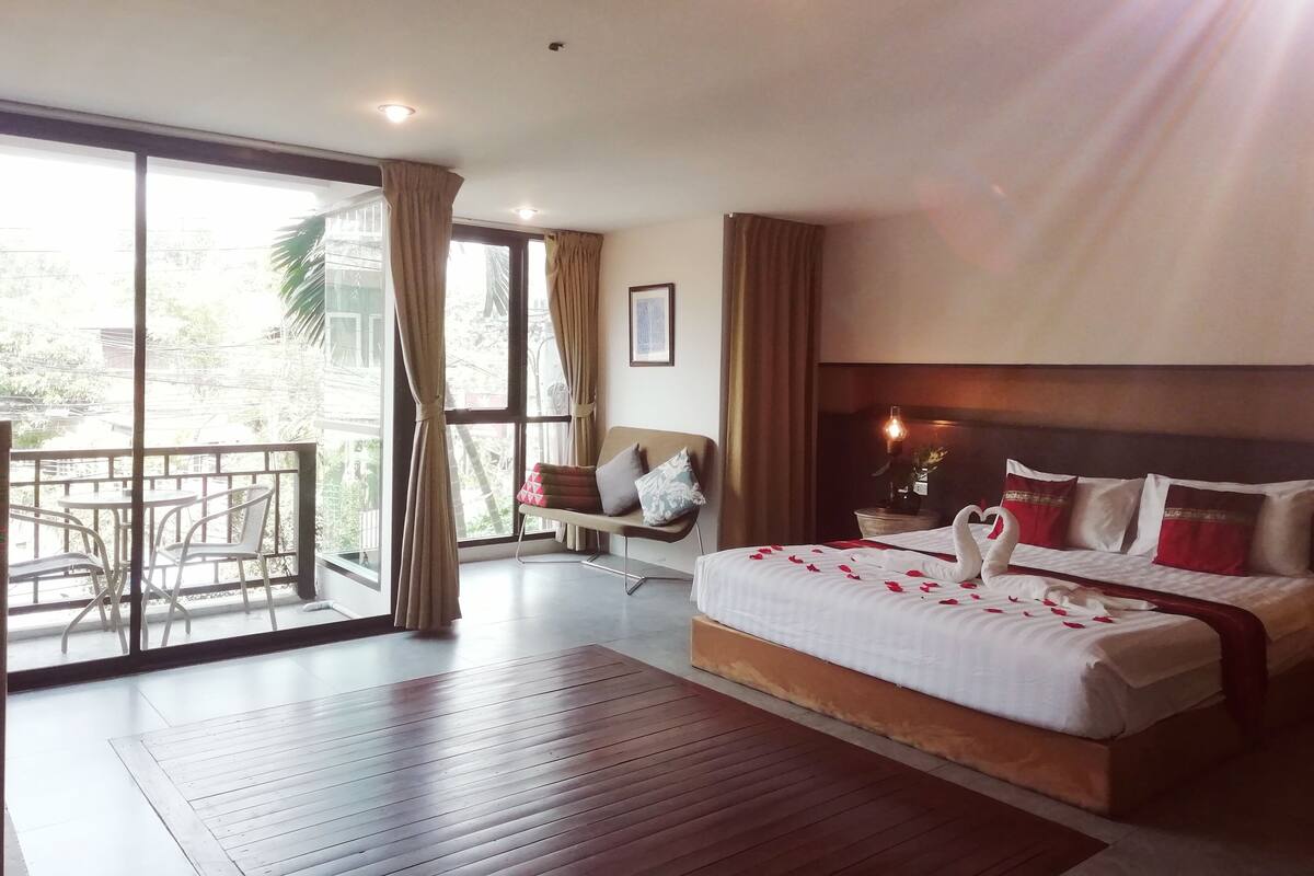 A hotel room with a large bed, a sofa, a balcony with a table and chairs, and a view of the outdoors.