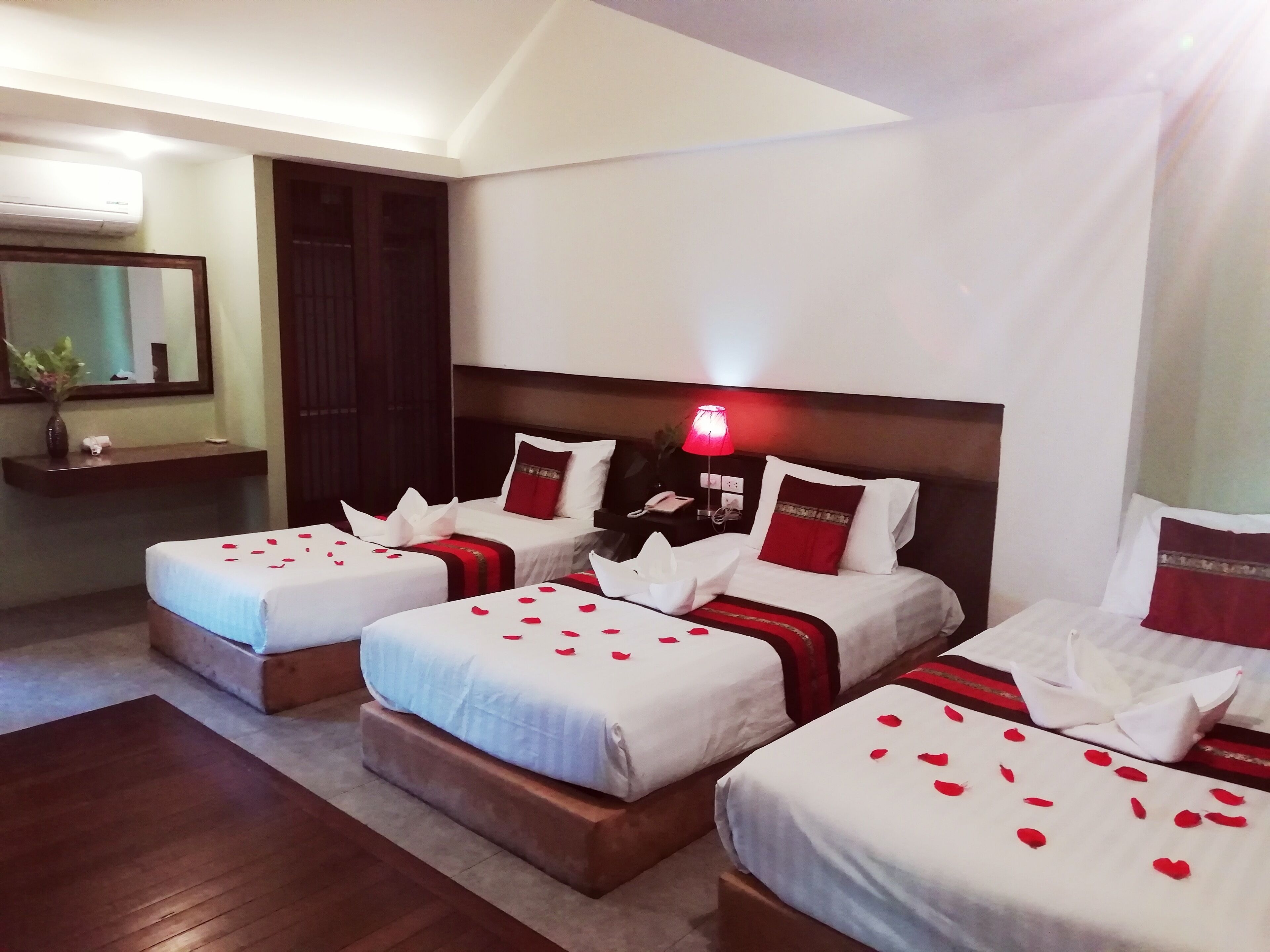 Habitación de hotel con tres camas, cada una con ropa de cama blanca y detalles rojos, una lámpara de noche y un espejo en la pared.