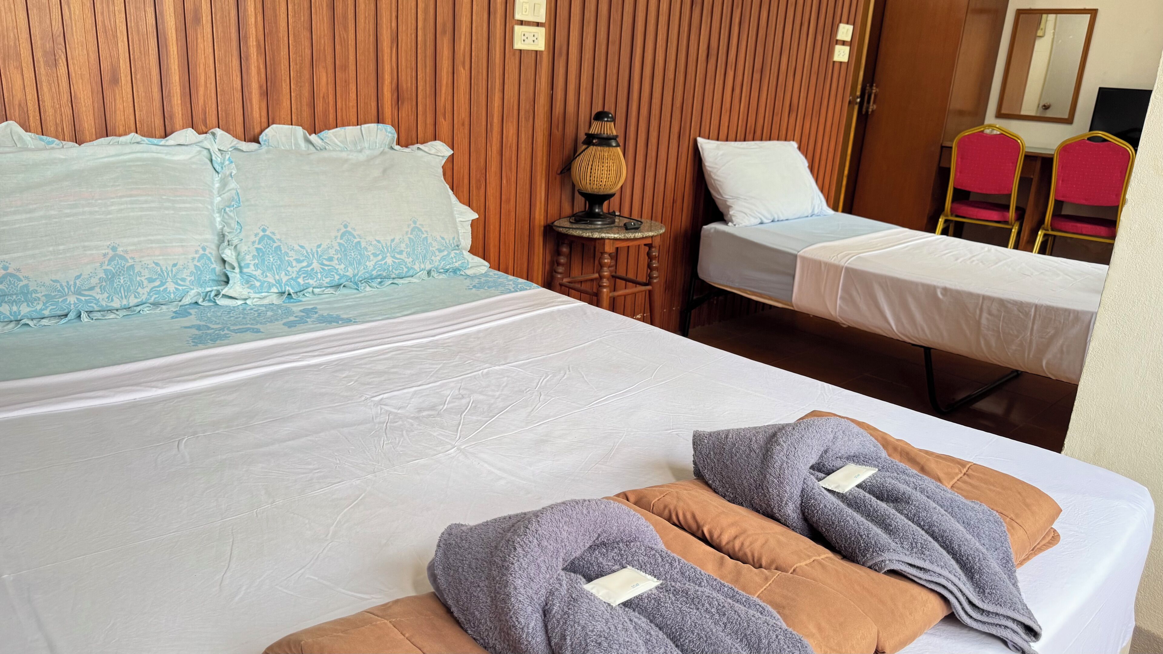 Hotellihuone, jossa on kaksi sänkyä, puinen seinä, pieni pöytä lampun kanssa ja peili.