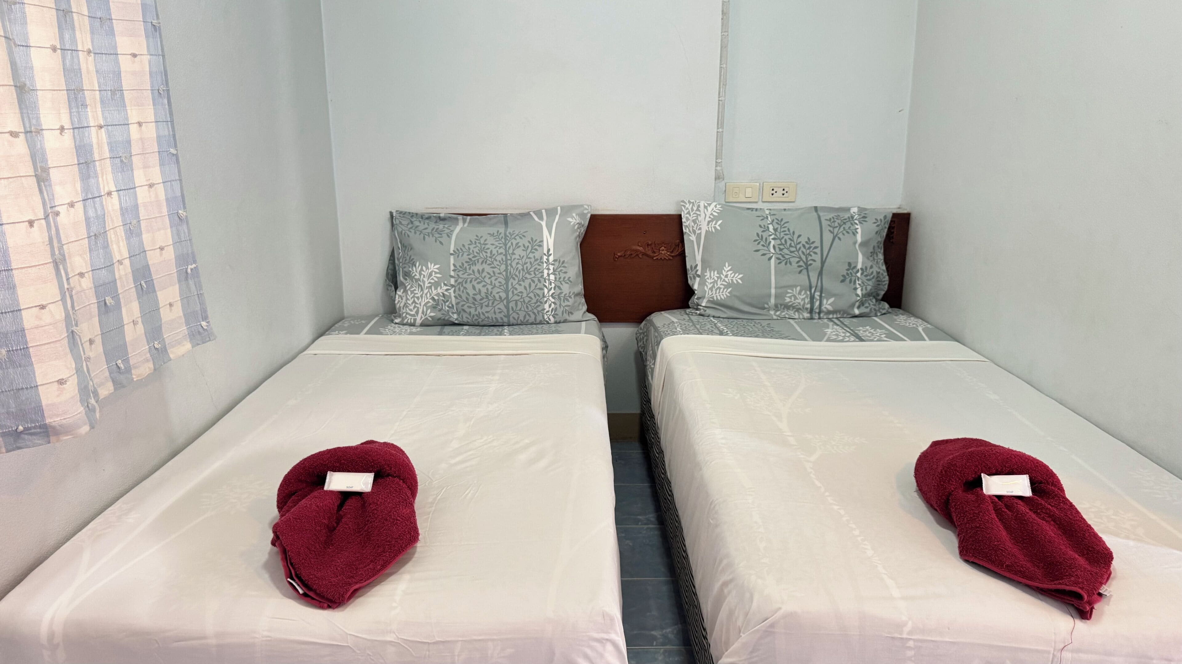 Two single beds with matching headboards and pillows, a window with a grid pattern, and a visible electrical outlet on the wall.