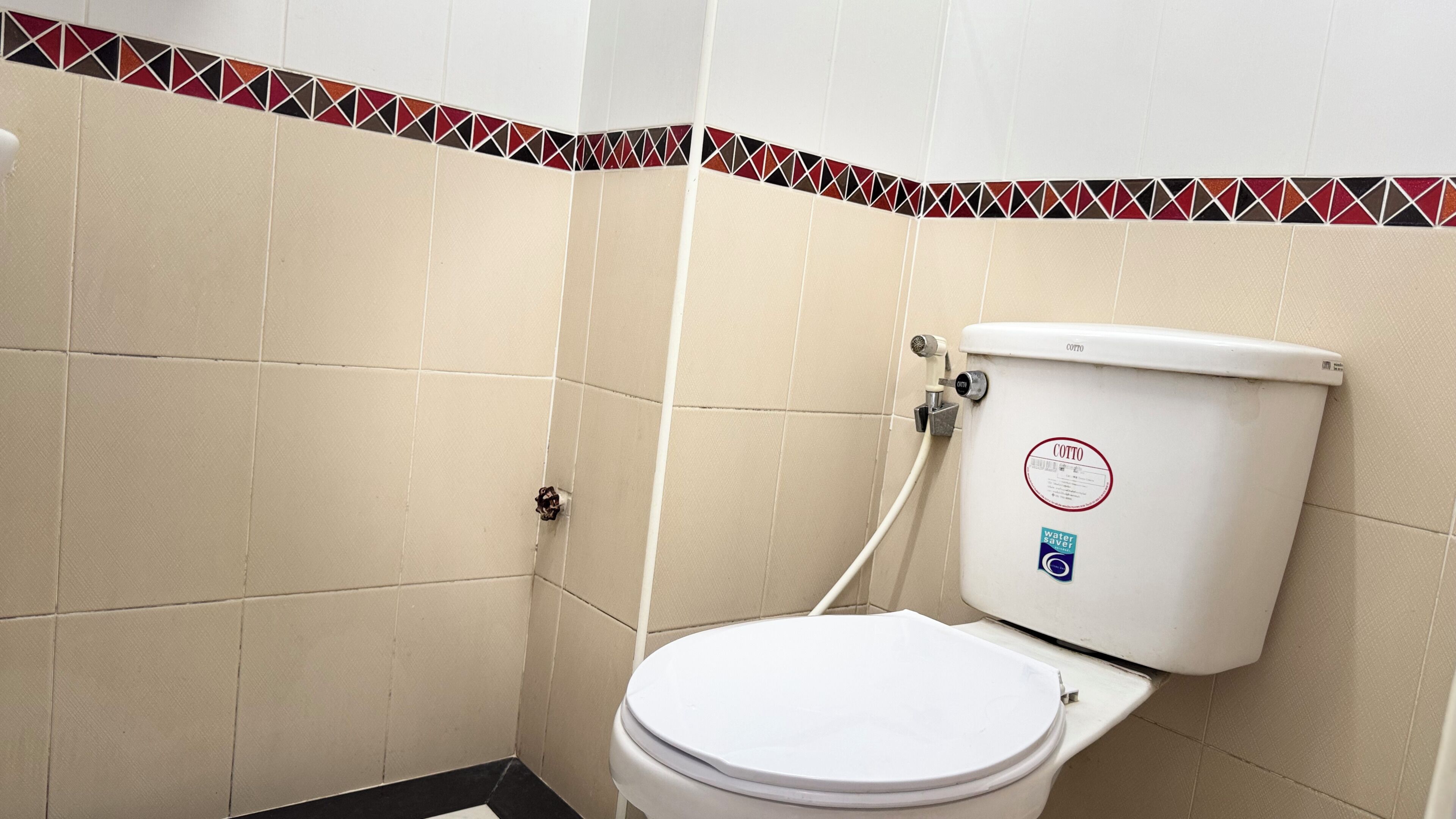 A small, tiled bathroom with a toilet, a water-saving sticker, and a red and black tile border.