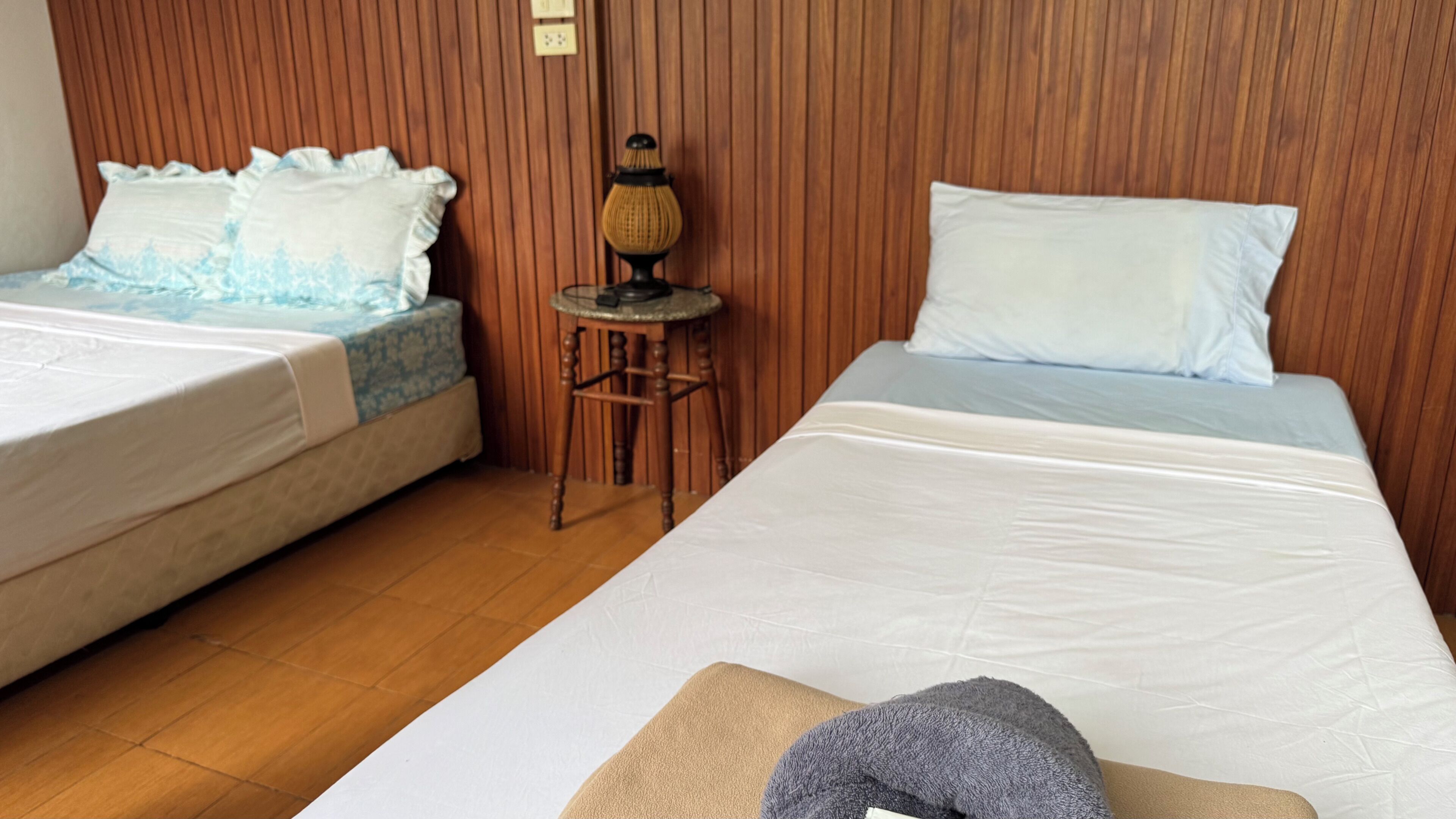 Two single beds with white linens, a wooden headboard, a small round table with a lamp, and a wooden floor.