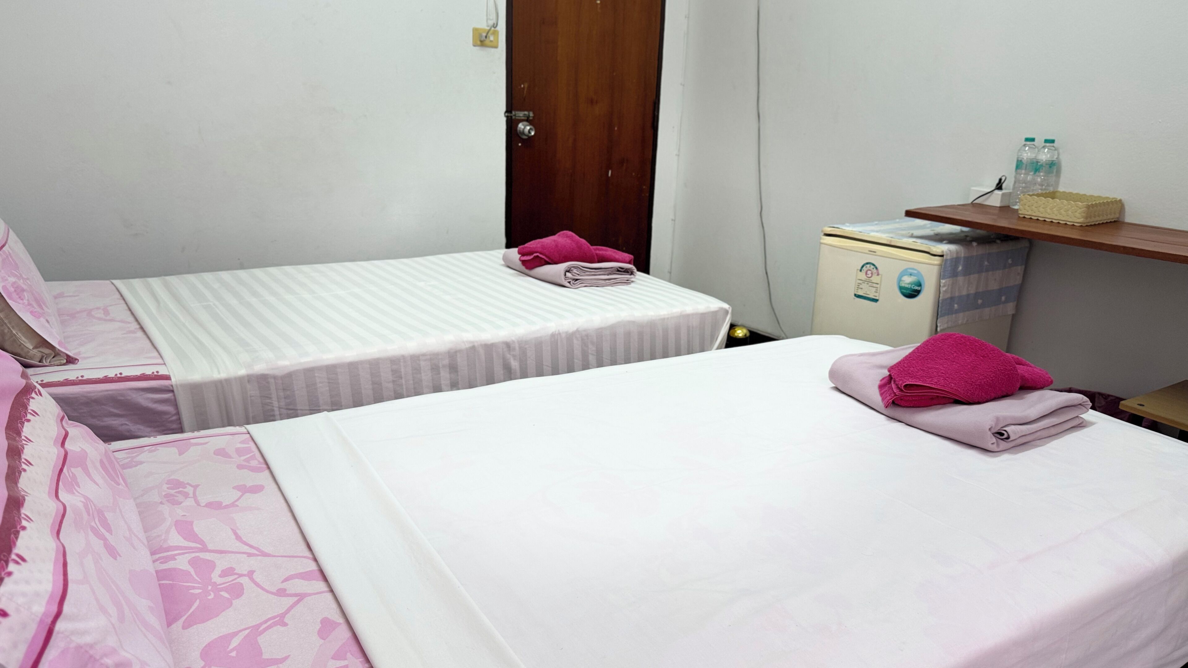 A room with two beds, a wooden door, a small refrigerator, and a shelf with bottles.