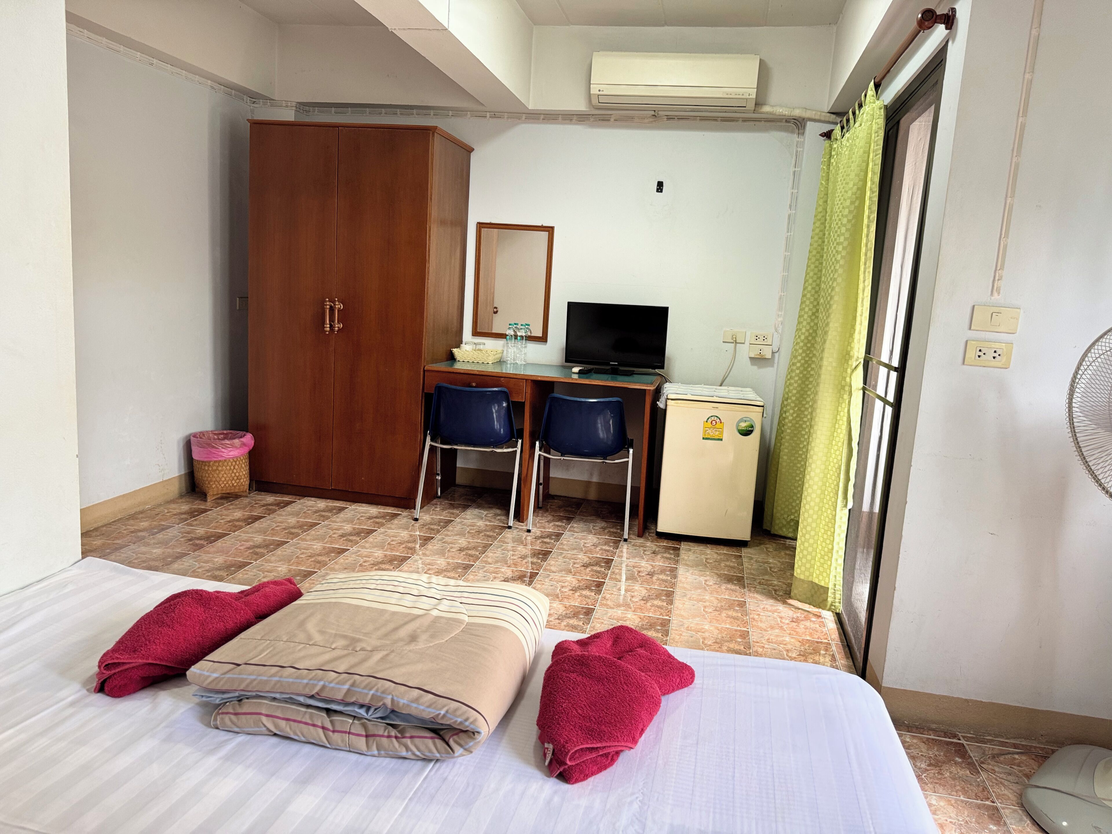 A hotel room with a bed, a desk with a computer, a wardrobe, a small fridge, and a fan.