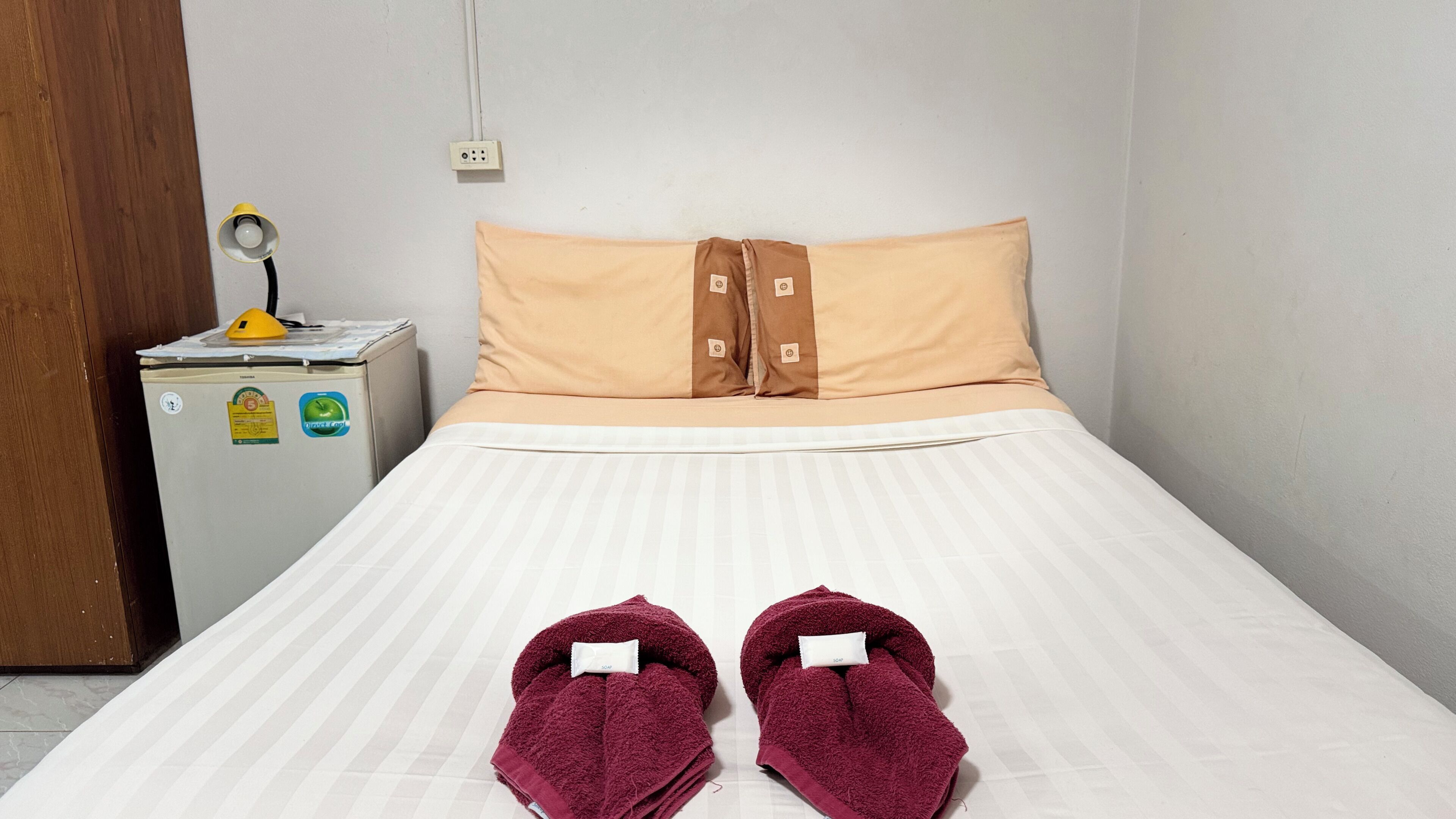 Un lit simple avec un matelas blanc et des oreillers beige, un petit réfrigérateur avec une lumière jaune et une paire de serviettes rouges posées sur le lit.