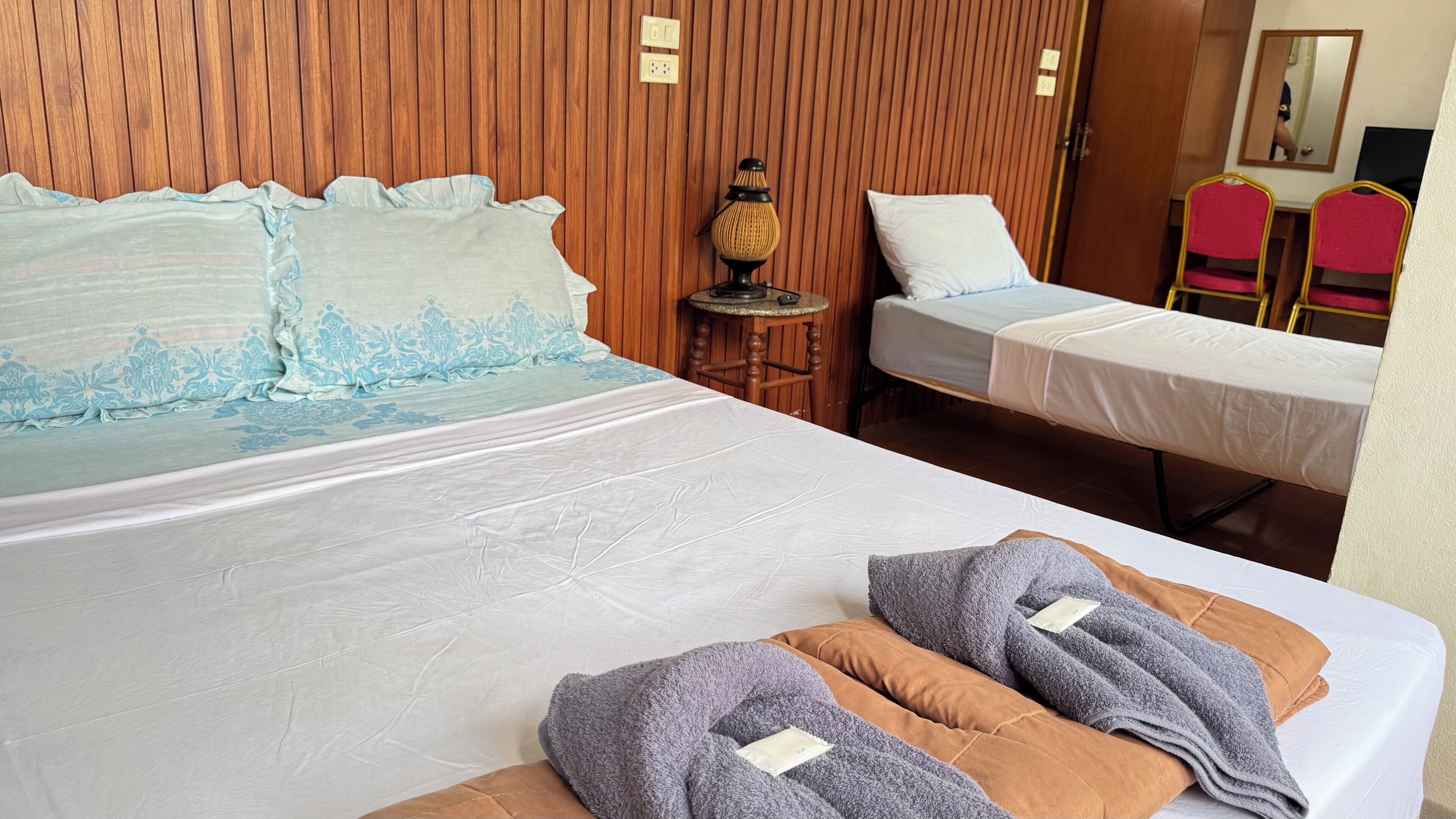 Quarto de hotel com uma cama grande, uma cama menor, cabeceira de madeira, uma mesinha com abajur e duas cadeiras.