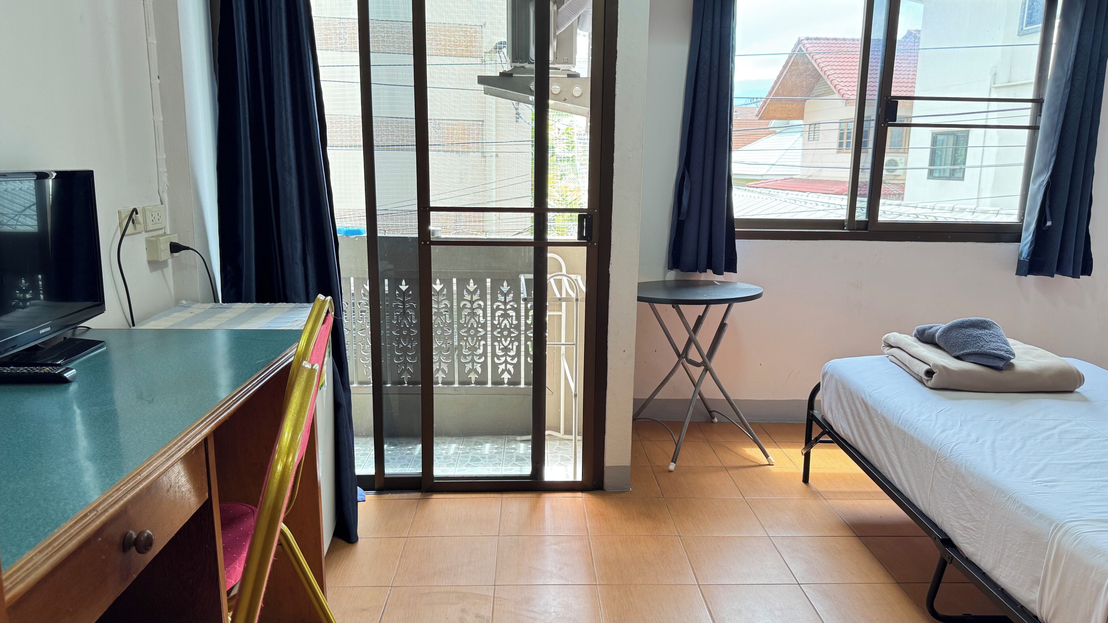 A room with a bed, a desk with a computer, a small table, and a view through a sliding door to an outdoor balcony.