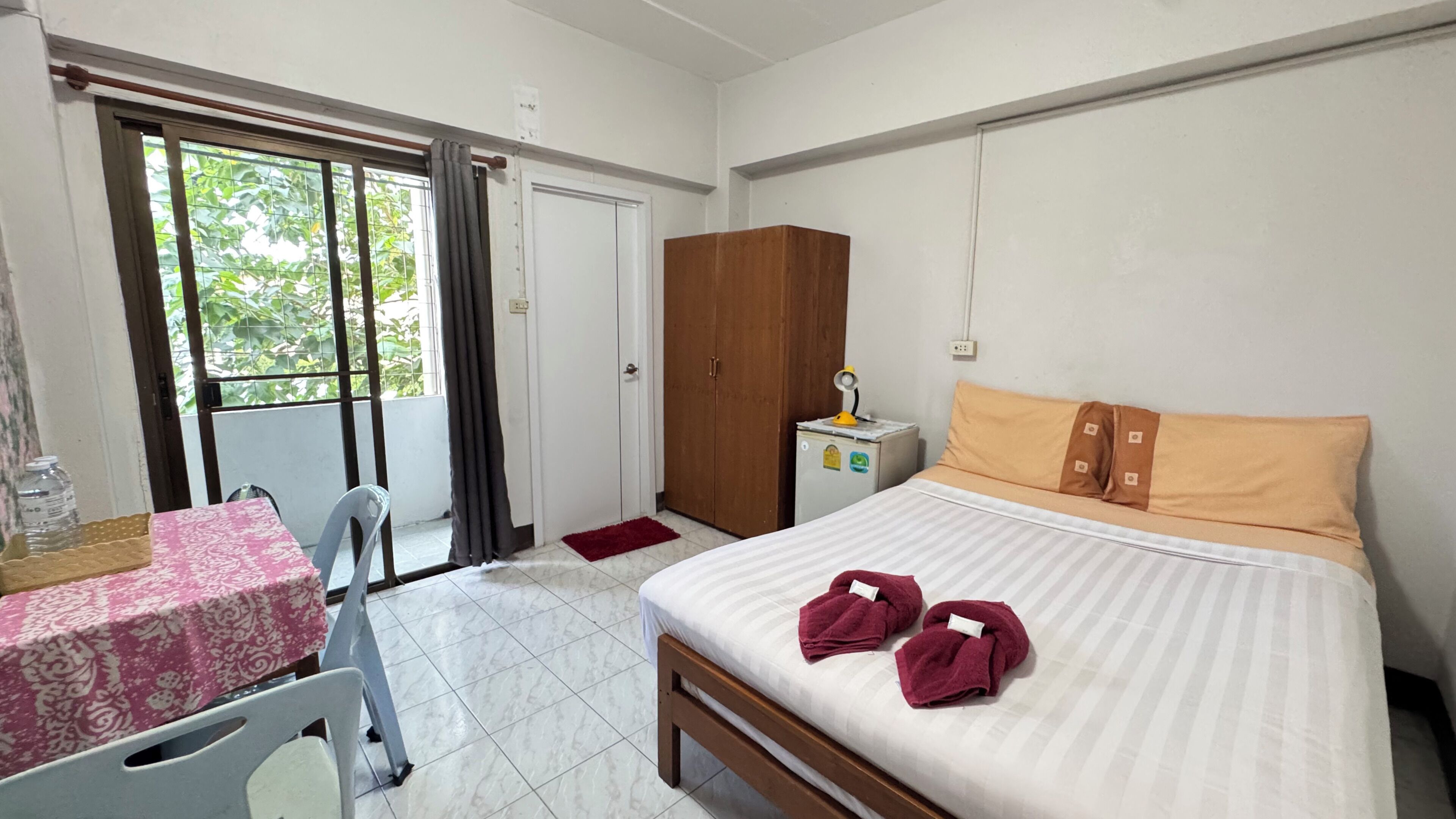A hotel room with a bed, a desk, a chair, a wardrobe, and a small table with a lamp.