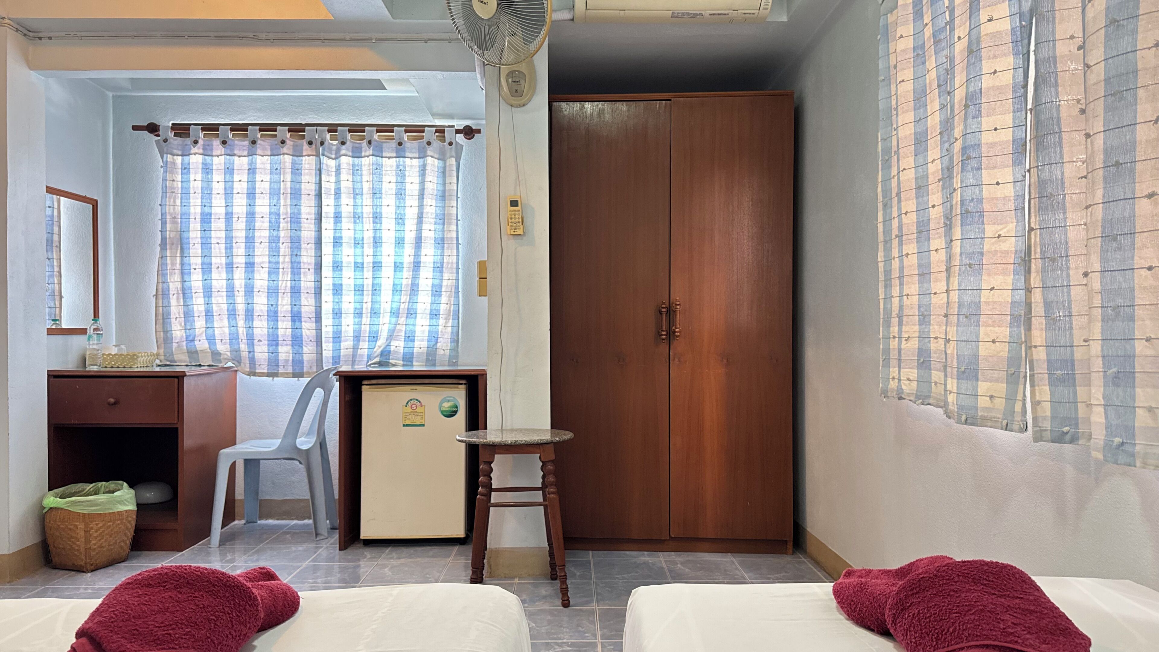 Una habitación con dos camas, un escritorio, un armario, una silla, un refrigerador pequeño y una ventana con cortinas.