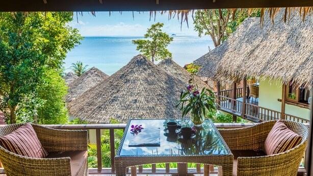 A tropical resort balcony with wicker furniture, a glass table, and a view of the ocean and thatched-roof huts. - Beyond The Blue Horizon Villa Resort (Ko Pha-ngan)
