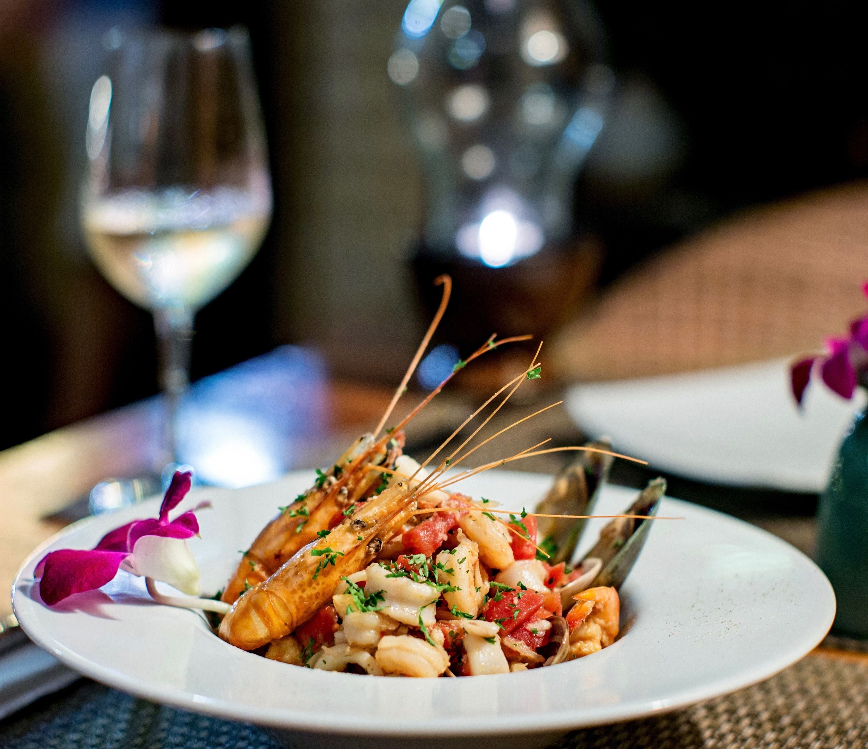 A plate of seafood with a glass of white wine and a vase with purple flowers on the table.